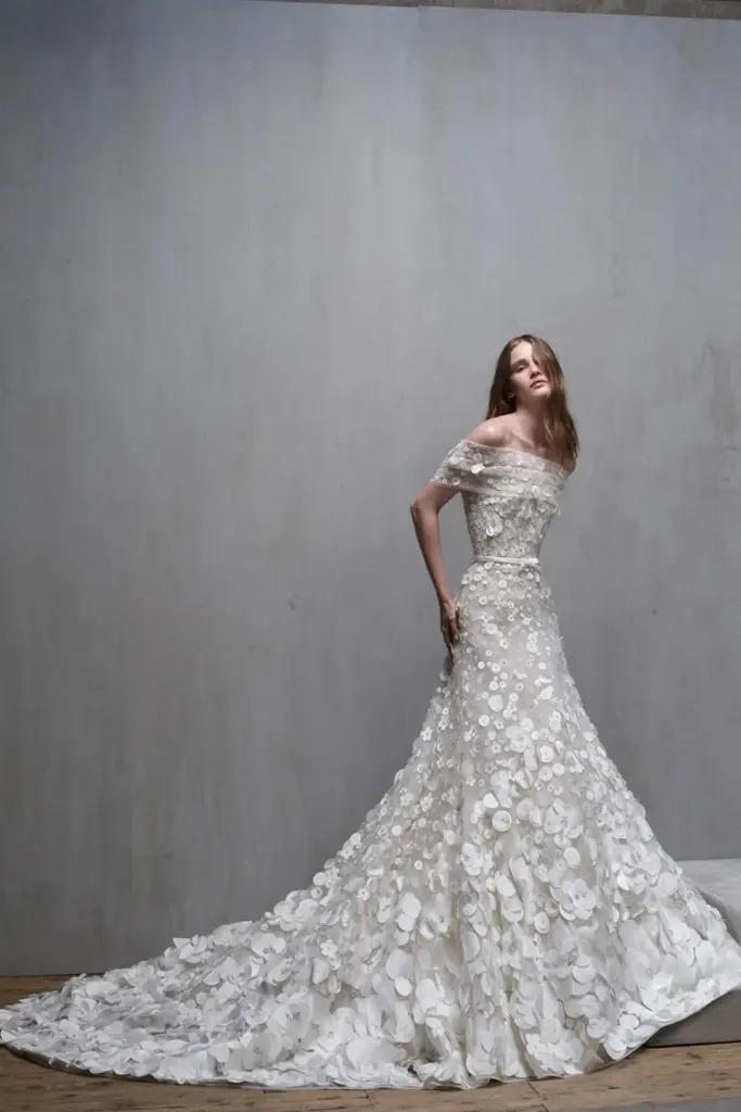 Elie Saab 2026 koleksiyonundan, üç boyutlu çiçek aplikeleriyle kaplanan off-shoulder gelinlik ve uzun kuyruk detayıyla romantik couture tasarım.
