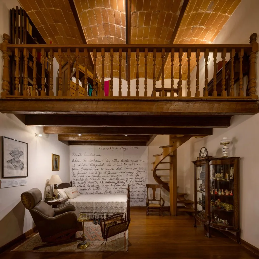 Museo Casa Kahlo’daki loftlu oda; ahşap merdiven, yatak, vitrin dolabı, Frida Kahlo’nun el yazısı duvar resmi ve sıcak ışıklandırmalı tarihi iç mekân tasarımı.