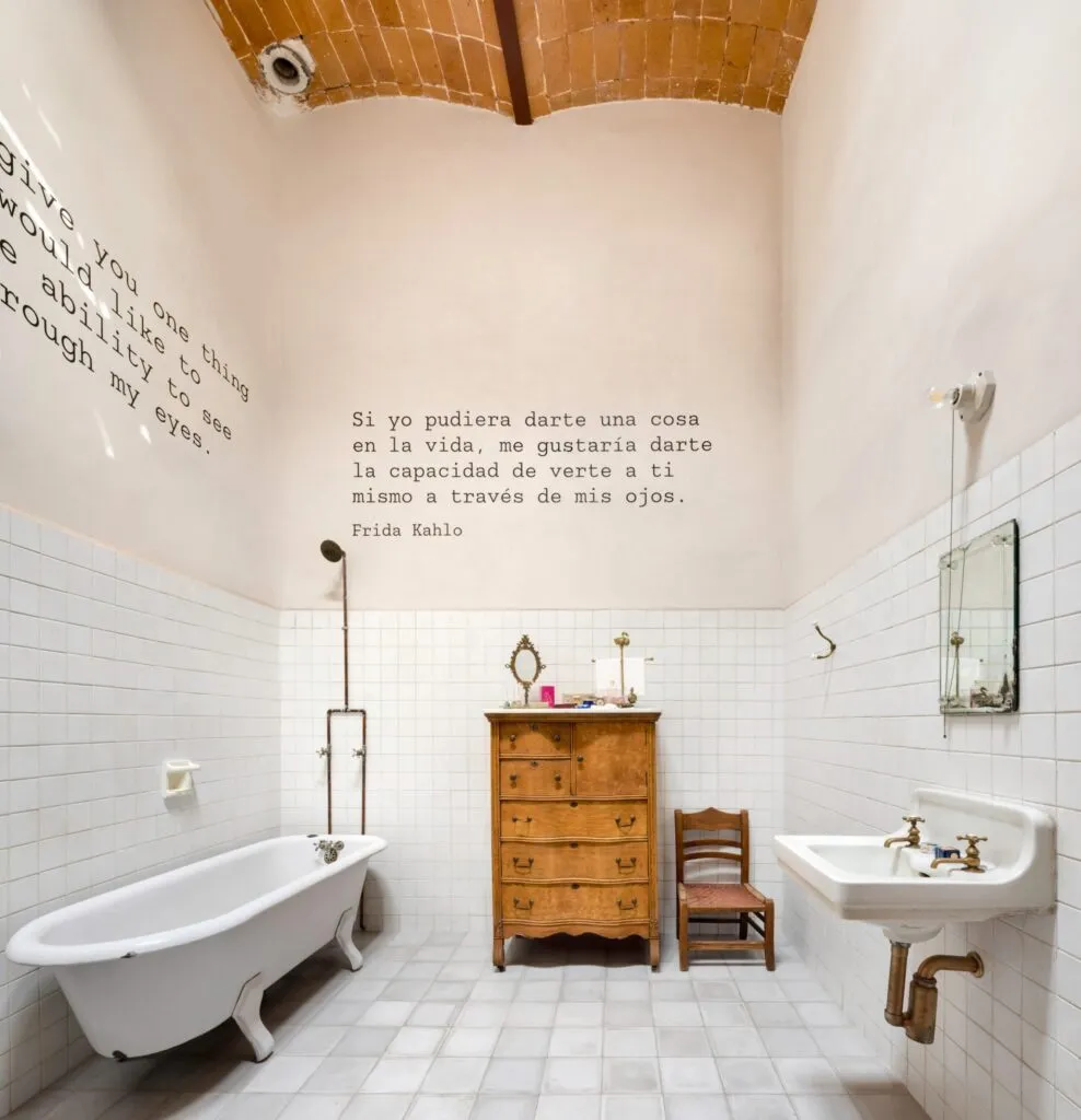 Frida Kahlo’nun aile evindeki banyo alanı; beyaz karo duvarlar, eski ahşap şifonyer, küvet ve duvarda Frida Kahlo’ya ait duvar yazılarıyla Museo Casa Kahlo’nun iç mekânı.