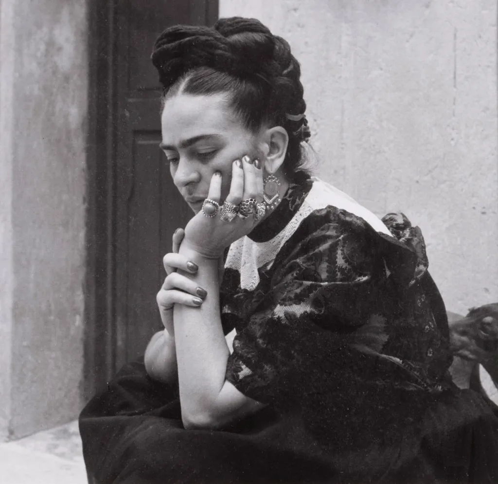 Siyah beyaz bu fotoğrafta, ikonik örgülü saçları ve yoğun kaşlarıyla tanınan Frida Kahlo, sağ elini düşünceli bir şekilde çenesine dayamış, parmaklarındaki gösterişli yüzükler ve ojeli tırnaklarıyla aşağıya doğru bakarak poz veriyor, arka planda sade bir duvar ve kapı görünüyor.