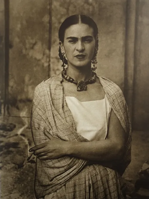 Frida Kahlo'nun kahverengi tonlardaki bu etkileyici portresinde, örgü saçları, birleşik kaşları, büyük küpeleri, kalın kolyesi ve desenli şalıyla ciddi bir ifadeyle kameraya bakan sanatçı, sağ elinde bir sigara tutuyor.