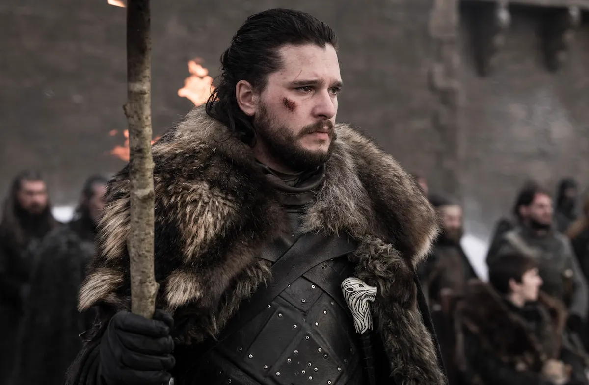 Yabancı diziler seçkisinde yer alan Game of Thrones dizisinde Jon Snow, kürklü pelerini ve kılıcıyla karanlık bir kale ortamında duruyor.
