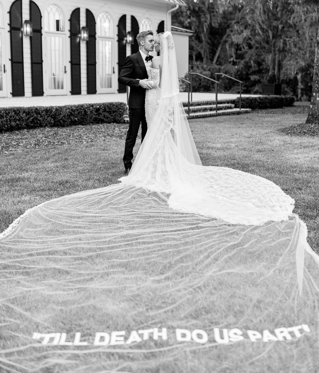 Uzun tül duvak üzerinde “Till Death Do Us Part” yazısı bulunan dantel detaylı gelinlik, modern ve ikonik düğün estetiğini yansıtan siyah beyaz çekim.