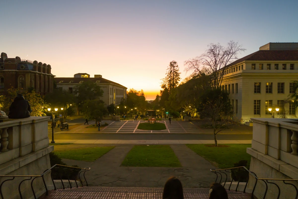 Kaliforniya Üniversitesi Berkeley kampüsünde, tarihi taş binalar ve ikonik saat kulesi Sather Tower'ın yemyeşil ağaçlar arasında yükseldiği, güneşli bir akademik ortamı gözler önüne seren geniş bir görünüm.
