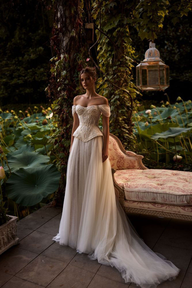 Doğayla iç içe romantik bir ortamda poz veren model, işlemeli korsajlı off-shoulder gelinlik ve tül etek detaylarıyla zarif bir peri masalı gelinlik görünümü sergiliyor.