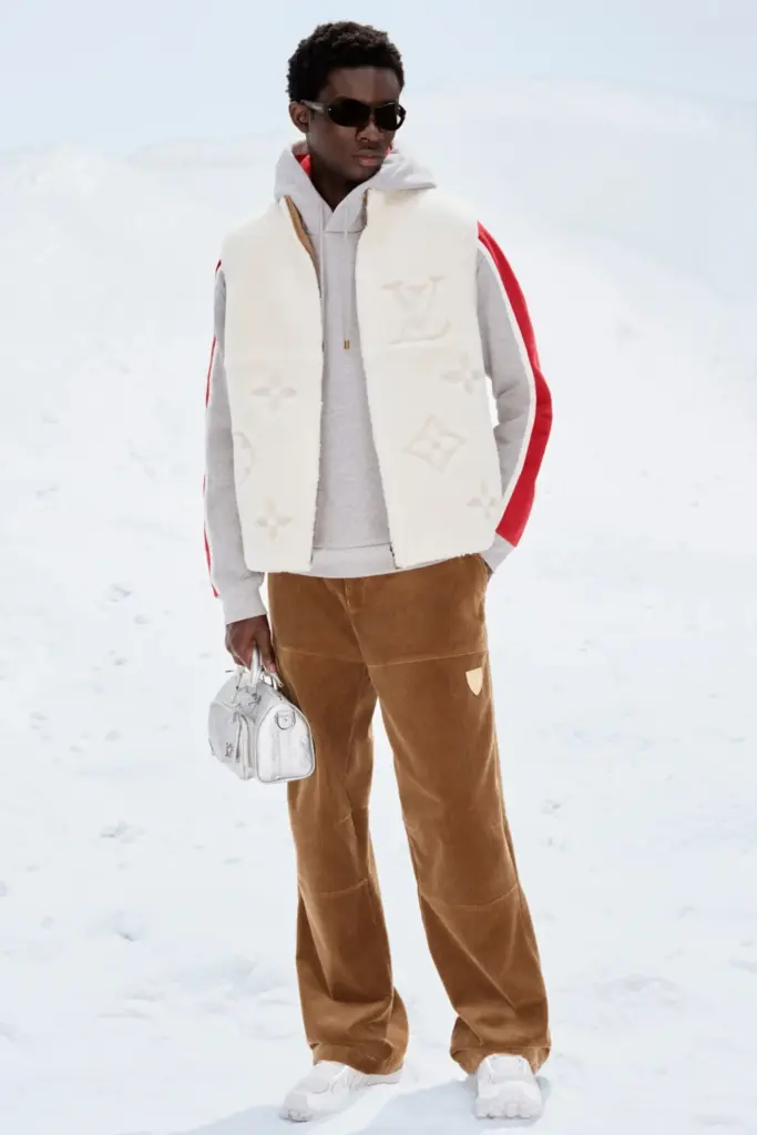 Louis Vuitton erkek kış kombini; beyaz monogram dokulu yelek, gri hoodie, taba pantolon ve mini gümüş el çantasıyla kar temalı modern lüks stil.