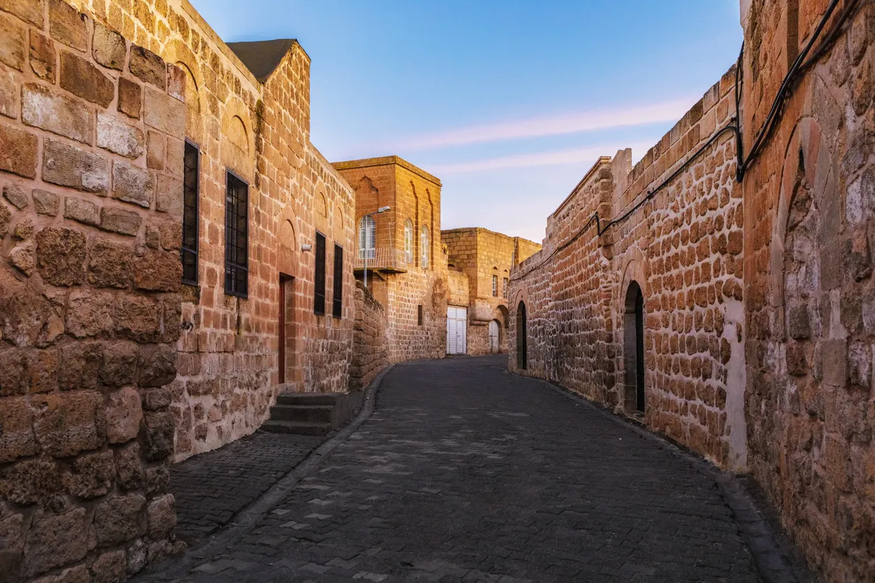 Mardin'in dar, eğimli, koyu renkli taş döşeli sokağında ilerlerken görünen, güneydoğuya özgü sarımsı taş bloklarla inşa edilmiş, pencereleri demir parmaklıklı ve bazıları kemerli girişlere sahip tarihi evler, gün batımının sıcak ışıklarıyla aydınlanıyor.