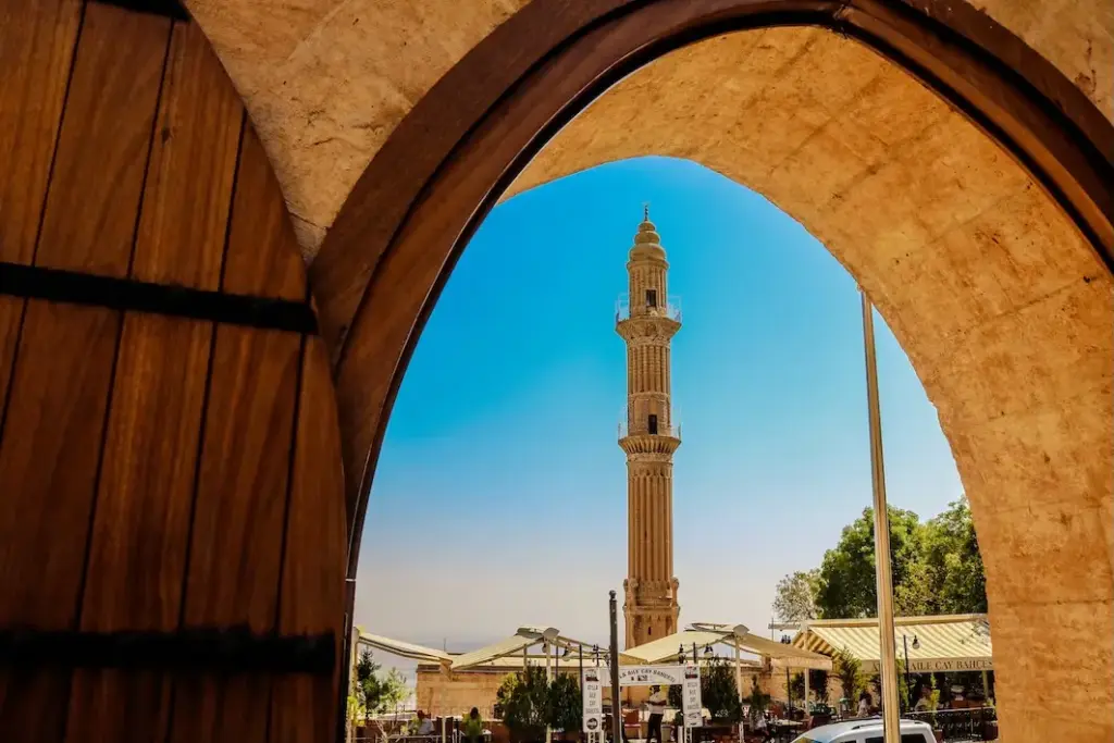 Sol taraftaki ahşap kapı ve kemerli taş bir geçitten görülen, masmavi gökyüzü altında yükselen, ince ve detaylı işlemeli Mardin Ulu Camii minaresi, altında yer alan hareketli çay bahçesiyle birlikte eşsiz bir mimari ve tarihi dokuyu gözler önüne seriyor.