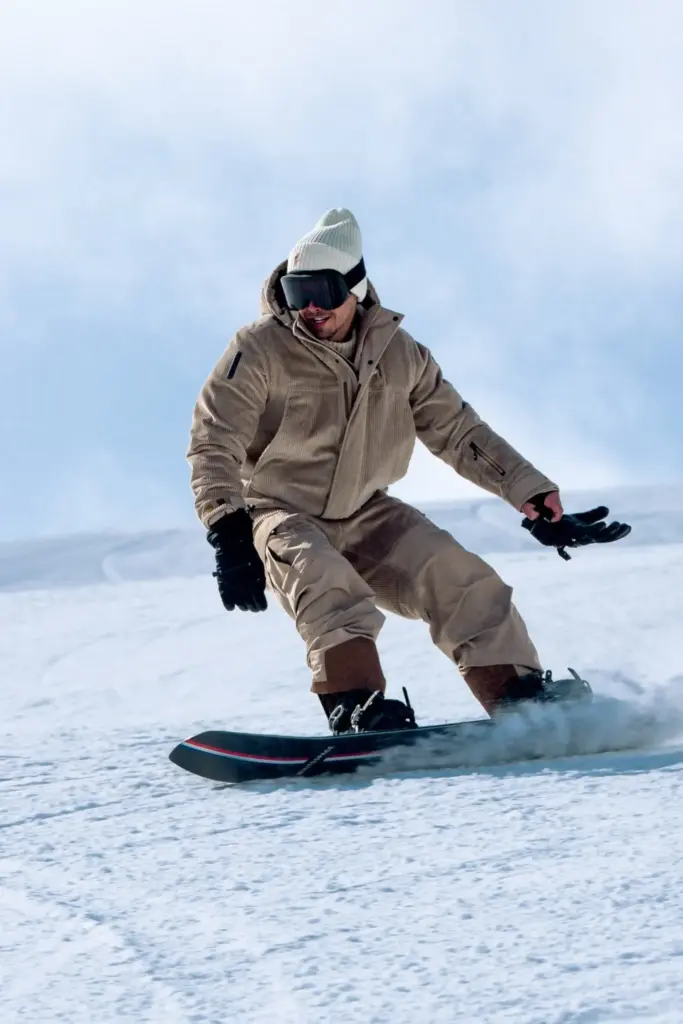 Karlı bir ortamda snowboard yapan ve üzerinde Moncler kayak koleksiyonundan devetüyü rengi kayak tulumu olan bir erkek