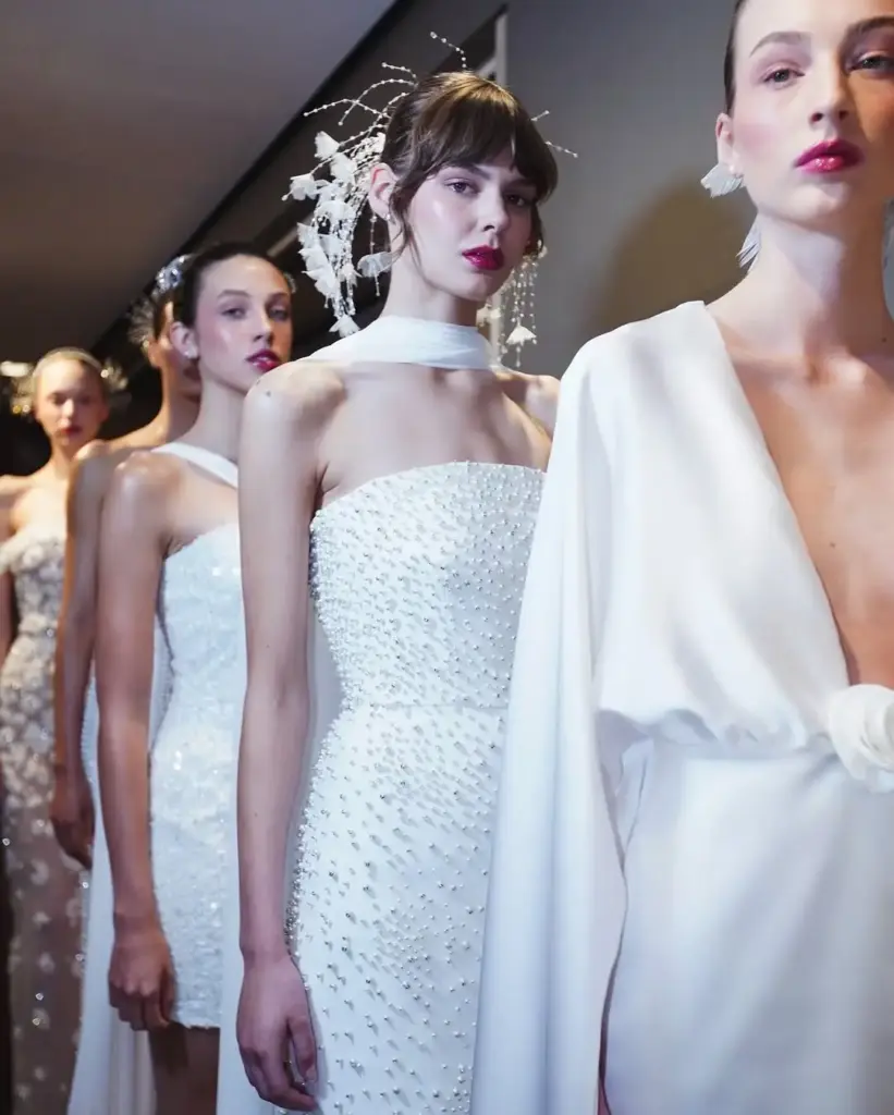 Backstage’de sıraya dizilmiş modeller, incili ve modern beyaz gelinlik tasarımlarıyla çekim öncesi bekliyor.