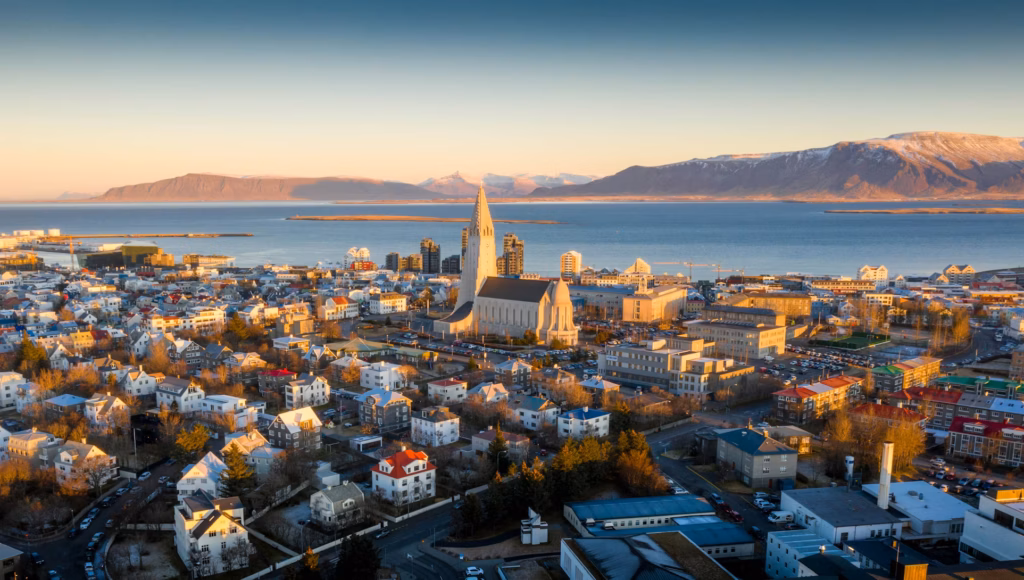 Reykjavik'in kuşbakışı görüntüsünde, altın sarısı gün batımı ışıkları altında yükselen ikonik Hallgrímskirkja Kilisesi, renkli çatılı evlerin arasından öne çıkarken, şehrin denize nazır konumu ve karlarla kaplı dağ manzarası İzlanda seyahat rehberi için büyüleyici bir genel bakış sunuyor.