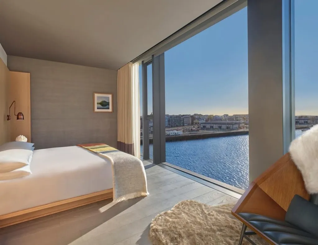 Modern bir otel odasının içinden, ahşap detaylar ve minimalist dekorasyonla döşenmiş bir yatağın ve kürk detaylı şık bir koltuğun bulunduğu alandan, zeminden tavana uzanan cam pencerelerle görünen masmavi bir marina ve Reykjavik şehir siluetinin gün ışığında nefes kesen manzarası.