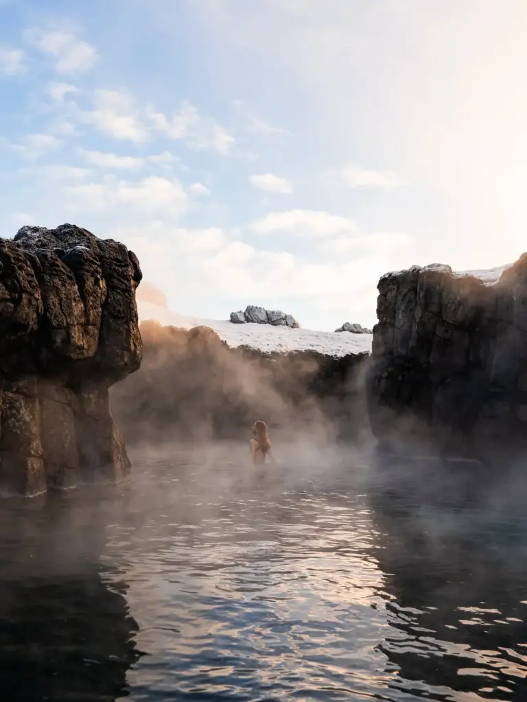 Reykjavik Seyahat Rehberi'nde bahsi geçen İzlanda'nın eşsiz doğal güzellikleri arasında yer alan, buharlar yükselen bir jeotermal havuzda koyu renkli kayalıkların arasında dinlenen bir kadın, güneşli ve bulutlu bir gökyüzünün altında huzurlu anlar yaşıyor.