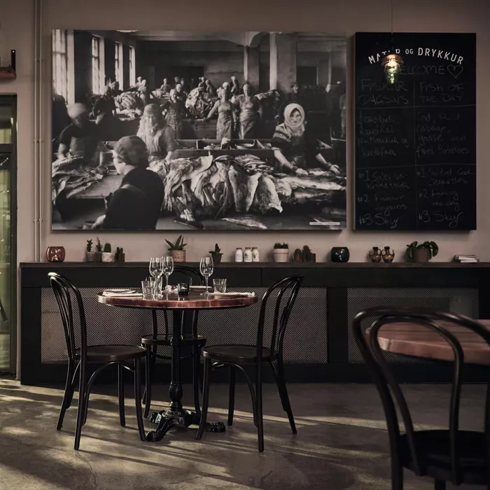 Reykjavik seyahat rehberi kapsamında öne çıkan bir İzlanda restoranının modern iç tasarımında, duvarda asılı, balık işleyen kadınları tasvir eden büyük siyah beyaz tarihi bir fotoğraf ile günlük menülerin yazıldığı tahta dikkat çekerken, zarifçe hazırlanmış masa ve sandalyeler misafirlerini bekliyor.