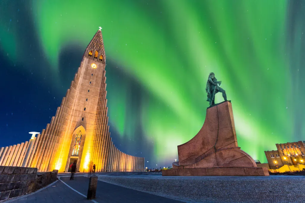 İzlanda'nın volkanik bazalt sütunlarından ilham alan mimarisiyle ikonik Hallgrímskirkja Kilisesi, ön planda Leif Erikson heykelinin bulunduğu meydanda, gece boyunca Kuzey Işıkları'nın büyüleyici yeşil parıltısı altında, Reykjavik seyahat rehberinin en unutulmaz manzaralarından birini sunuyor.