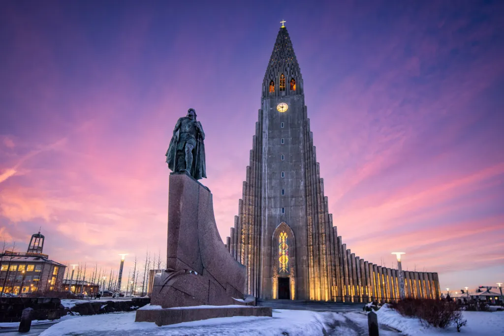 Mor ve pembe tonlarında gün batımı gökyüzünün altında karlı bir zeminde yükselen, bazalt sütunlardan ilham alan Hallgrímskirkja Kilisesi ve önündeki Leif Erikson heykeli, Reykjavik seyahat rehberi için etkileyici bir kış manzarası sunuyor.