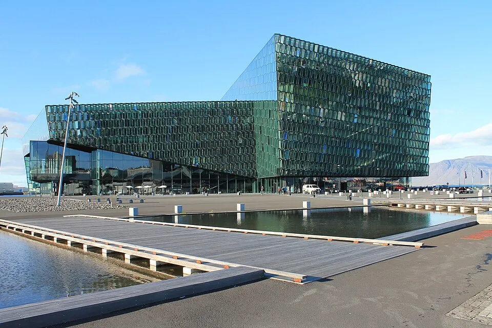Reykjavik'in ikonik yapılarından Harpa Konser Salonu, camdan petek cephesi ve önündeki sakin su havuzlarıyla modern mimarinin etkileyici bir örneğini sunarken, uzaklardaki dağlar şehrin doğal güzelliğini vurguluyor.