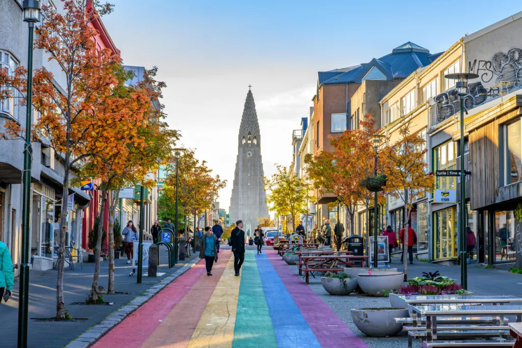 İzlanda'nın başkenti Reykjavik'in kalbinde, Hallgrímskirkja kilisesine doğru uzanan, gökkuşağı renkleriyle boyanmış meşhur Rainbow Street üzerinde sonbahar yapraklı ağaçlar arasında yürüyen insanları ve hareketli şehir yaşamını gösteren bir kare.