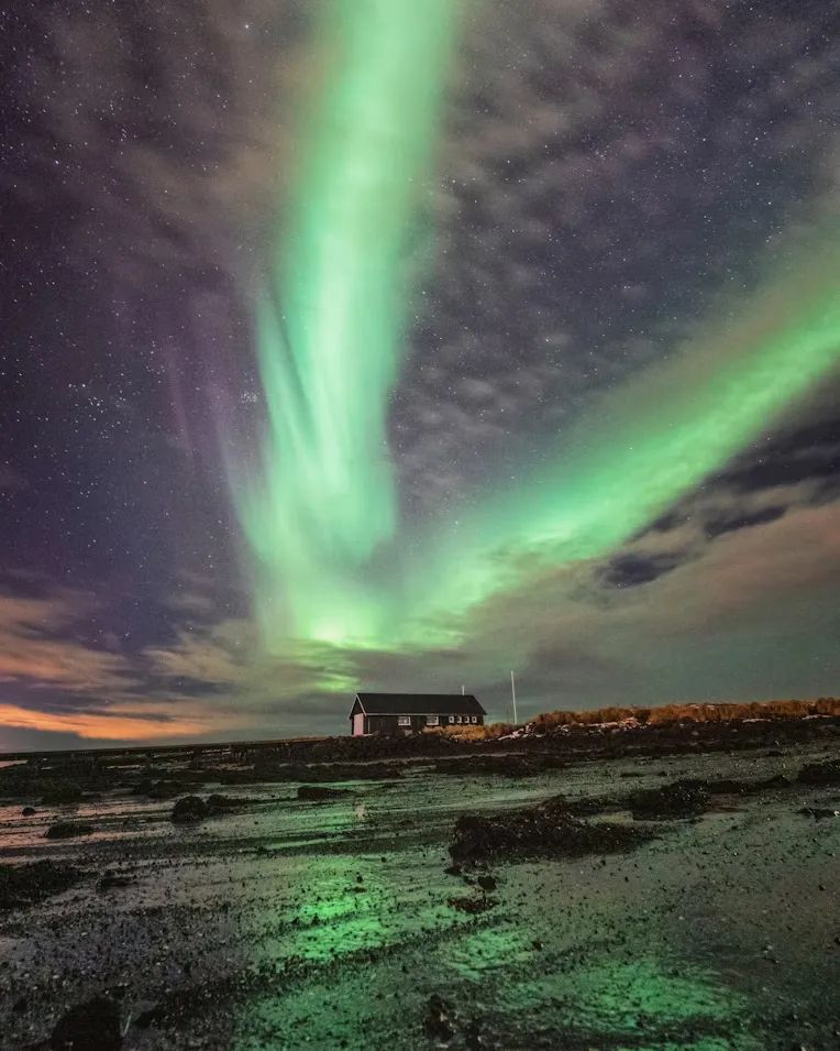 Reykjavik seyahati için önemli bir deneyim olan Kuzey Işıkları'nın, yıldızlarla dolu karanlık gökyüzünde göz alıcı yeşil huzmelerle dans ederek, ıslak ve kayalık zeminin üzerinde yalnız duran küçük bir evin bulunduğu ıssız bir manzarayı aydınlattığı nefes kesici bir an.