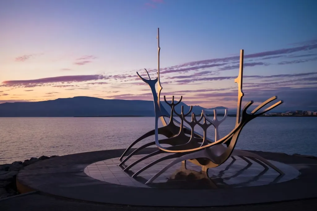 Gün batımının mor, pembe ve turuncu tonlarıyla ışıldayan gökyüzü altında, Reykjavik'in deniz kıyısında yer alan, Viking gemisi iskeletini andıran modern çelik Sun Voyager heykeli, İzlanda'nın keşif ve yolculuk ruhunu yansıtan bu özel seyahat rehberi için büyüleyici bir açılış sunuyor.