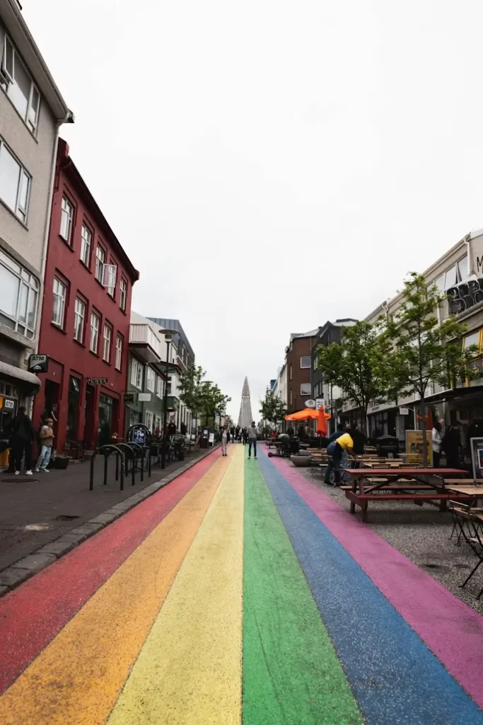 Reykjavik seyahat rehberi kapsamında, şehrin ikonik Rainbow Street'i üzerinde, uzakta görünen Hallgrímskirkja Kilisesi'ne doğru ilerleyen, gökkuşağı renkleriyle boyanmış hareketli bir cadde boyunca yürüyen insanlar, yol kenarındaki kafelerin masaları ve renkli binalar, bu canlı ve sanatsal atmosferi vurguluyor.