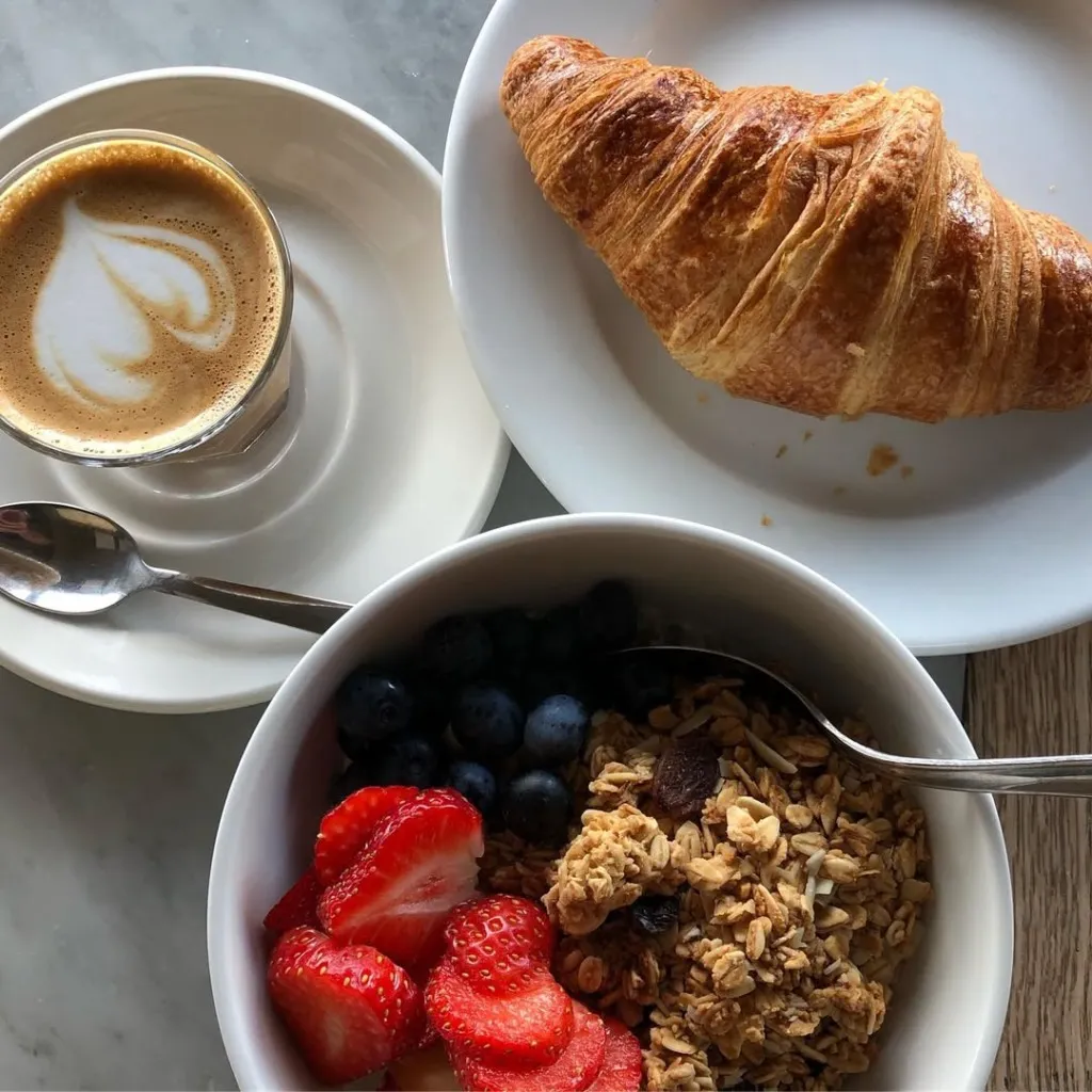 Masada beyaz bir tabağın üzerinde altın rengi çıtır bir kruvasan, yanında kalpli latte art'lı bir kahve ve çilek, yaban mersini ile granolalı sağlıklı bir kahvaltı kasesi Reykjavik Seyahat Rehberi'nde keşfedilebilecek kafe deneyimlerinin lezzetli bir örneğini sunuyor.