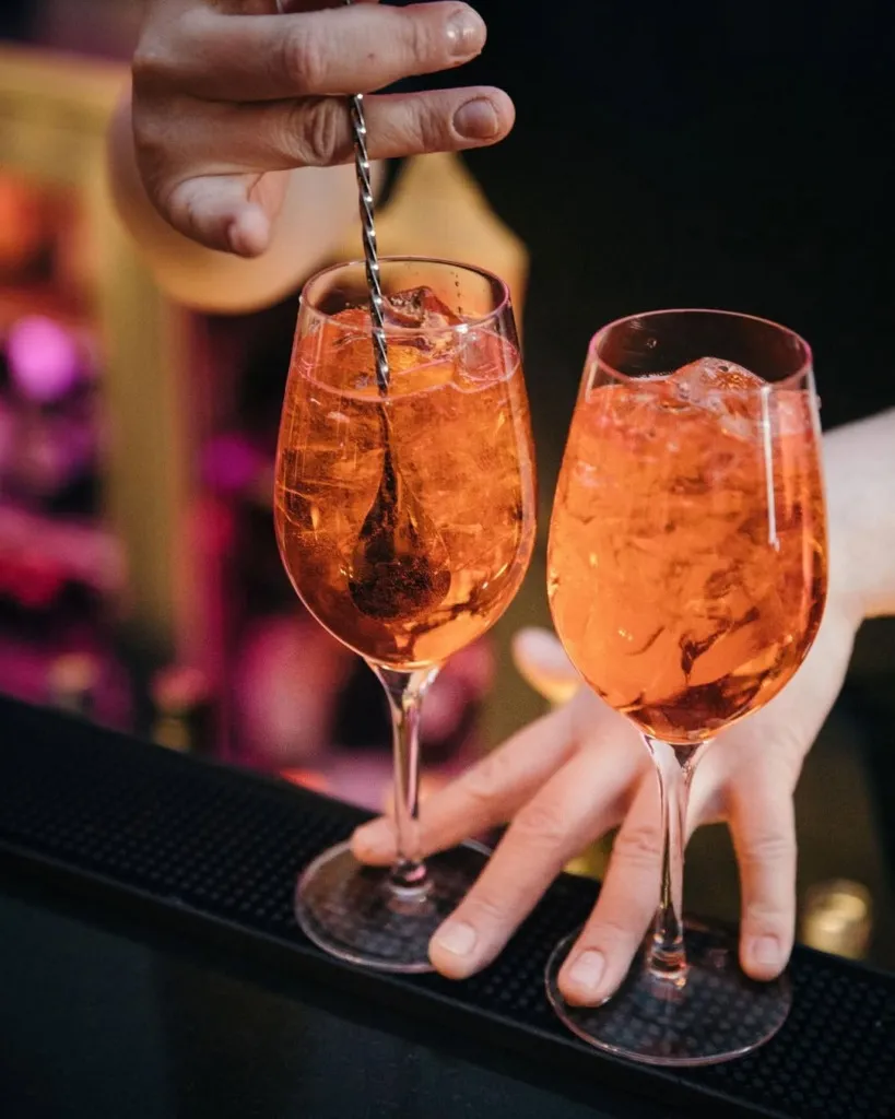 Reykjavik seyahat rehberi kapsamında şehrin canlı akşam atmosferini ve keyifli kokteyl duraklarını keşfetmeye davet eden bu görselde, loş ışıklı bir bar ortamında, barmen iki adet buzlu, turuncu renkli kokteylden birini uzun bir karıştırma kaşığıyla dikkatlice karıştırırken görülüyor.