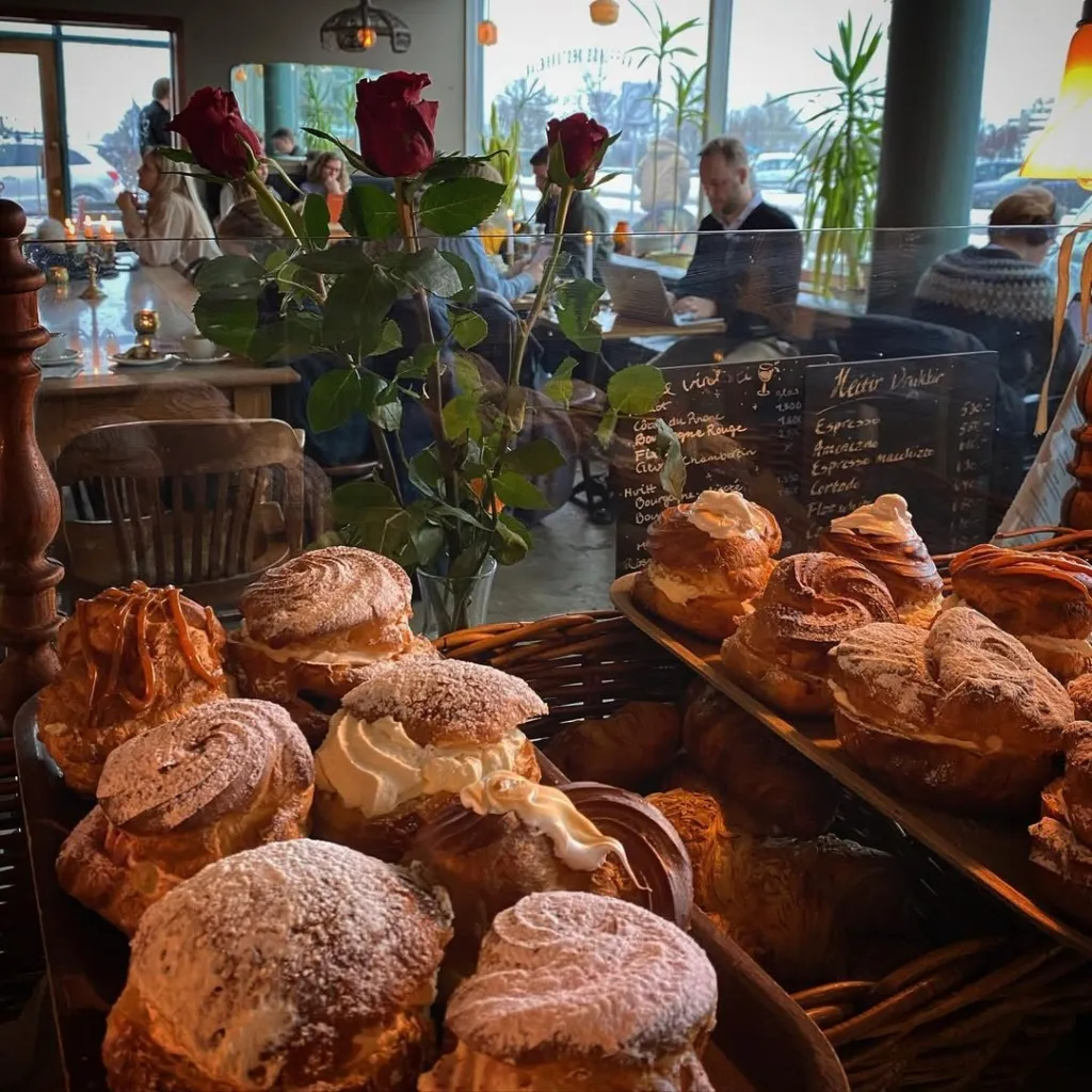 Reykjavik'teki şık bir fırın veya kafenin vitrininde, pudra şekeriyle süslenmiş bol kremalı tatlılar, karamelli kruvasanlar ve taze hamur işleri iştah açıcı bir şekilde sergilenirken, arka planda oturan konuklar ve kırmızı güllerin yer aldığı masalar şehrin bu özel duraklarını keşfetmek isteyenler için keyifli bir Reykjavik seyahat rehberi deneyimi sunuyor.