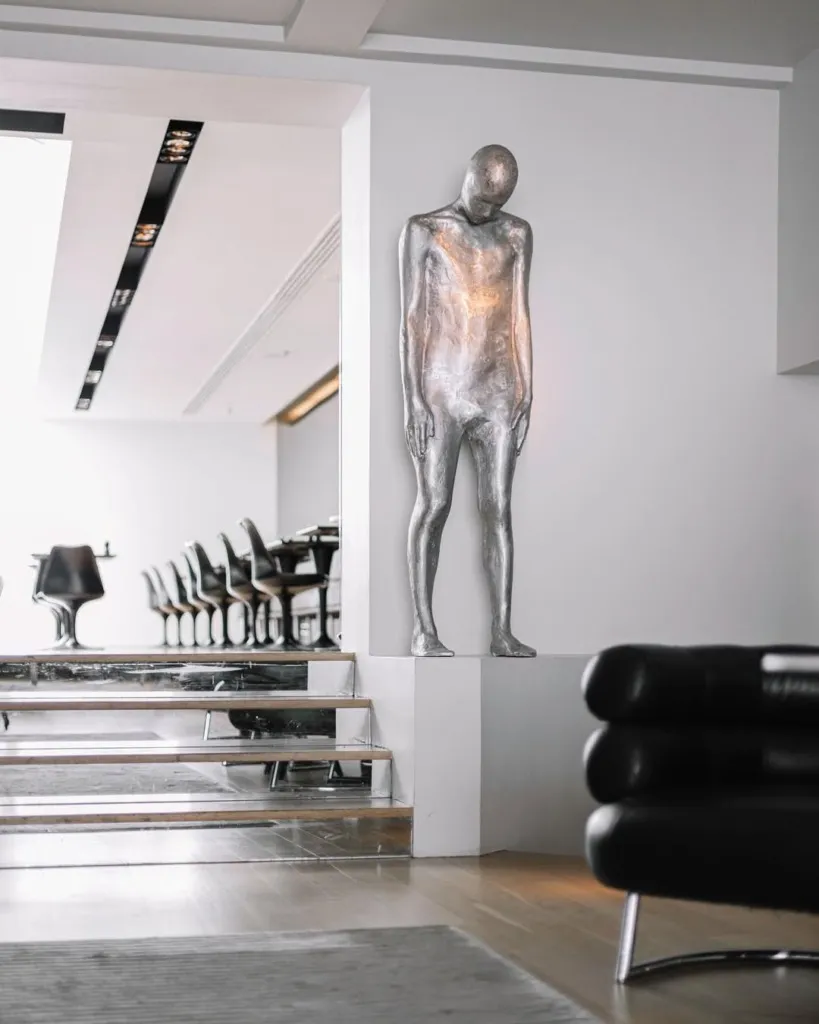 Reykjavik'in tasarım odaklı modern bir otelinde, beyaz bir duvarın önünde duran, çağdaş sanatın minimalist bir yorumu olan gümüş renkli, insan figürlü heykel, otelin sofistike ve sanatsal atmosferini vurguluyor.