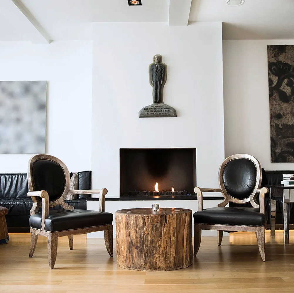 Minimalist çizgilerle dekore edilmiş modern bir Reykjavik otel lobisinde, şöminenin önünde yer alan iki klasik siyah deri koltuk, doğal ağaç kütüğünden yapılmış sehpayla sofistike bir atmosfer oluştururken, çağdaş sanat eseri detayıyla şehrin estetik ruhunu yansıtıyor.