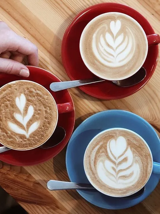 Aydınlık ahşap bir masanın üzerinde, zarif yaprak motifli latte art'larla süslenmiş, biri mavi ikisi kırmızı fincan tabağında duran üç sıcak kahve bardağı ve bunlardan birine uzanan bir el, Reykjavik seyahat rehberi kapsamında yer alan şehirdeki kafelerden keyifli bir anı yansıtıyor.