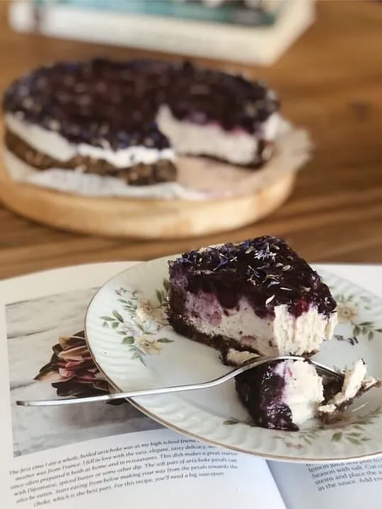 Çiçek desenli bir tabakta duran, üzeri koyu mor yaban mersini soslu ve kuru çiçeklerle süslü, bir çatalla yarısı yenmiş kremalı bir cheesecake dilimi, arkada ahşap tepsideki bütün pastanın bulanık görüntüsüyle, Reykjavik seyahat rehberinin sıcak kafe önerileri atmosferini yansıtıyor.