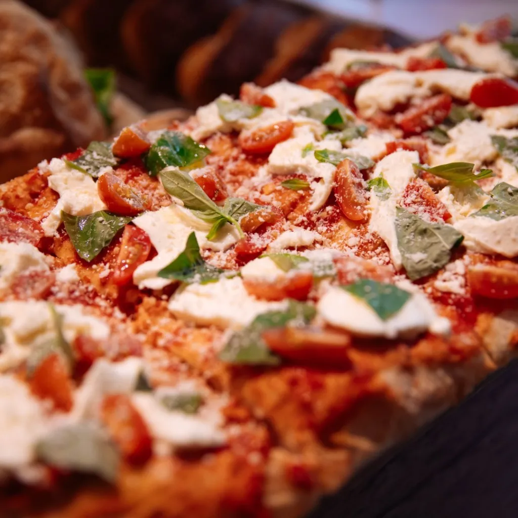 Reykjavik seyahat rehberi içeriğindeki yerel lezzet duraklarından, taze domates sosu, mozzarella peyniri, dilimlenmiş cherry domatesler ve fesleğen yapraklarıyla zenginleştirilmiş, üzerine bolca rendelenmiş peynir serpilmiş iştah açıcı bir fırın pizzası veya pide, şehirdeki kafe ve fırınların sunduğu hafif öğle yemeği seçeneklerini vurguluyor.