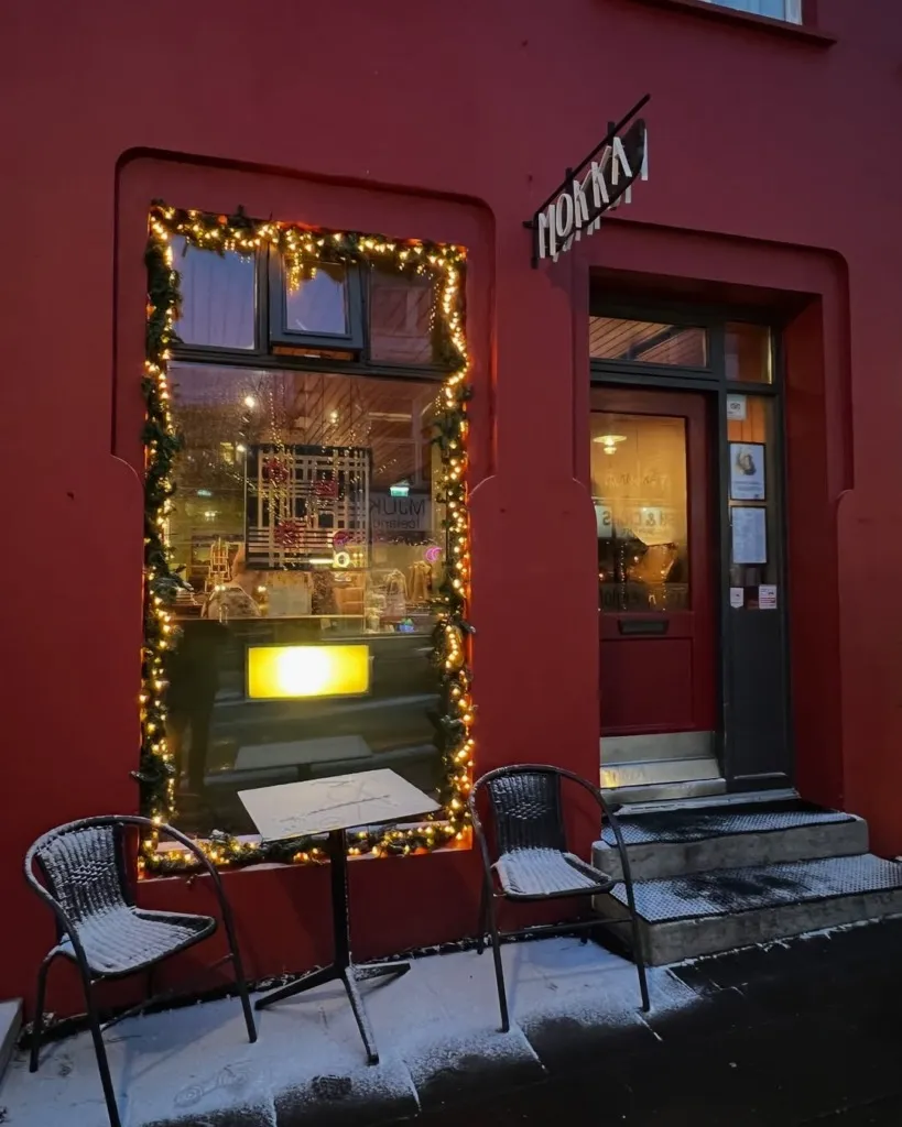 Kırmızı bir binanın ön cephesinde, vitrini sıcak ışıklarla ve yeşil çelenklerle süslenmiş Mokka Kaffi, dışarıda hafifçe karla kaplı masa ve sandalyeleriyle Reykjavik seyahat rehberindeki klasik kafelerden birinin davetkar kış atmosferini sunuyor.