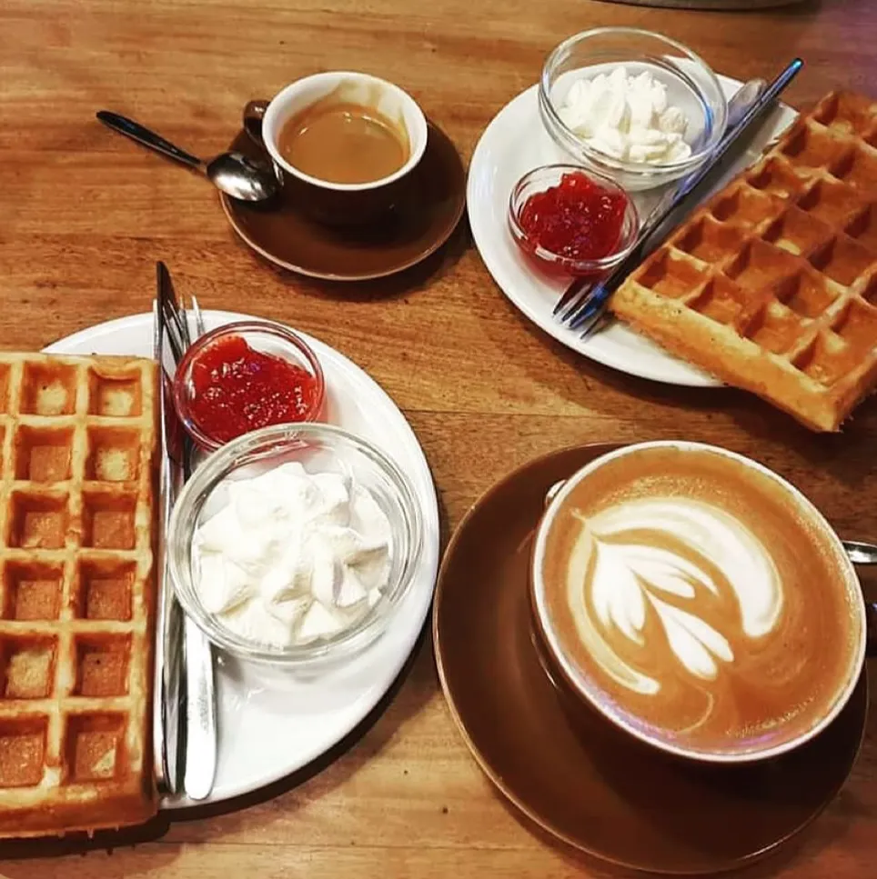 Tahta bir masanın üzerinde, yanında küçük cam kaselerde kırmızı reçel ve çırpılmış krema ile servis edilmiş iki porsiyon waffle ile birinde latte sanatı bulunan iki fincan kahve, sıcak bir Reykjavik kafesinde keyifli bir seyahat anını yansıtıyor.