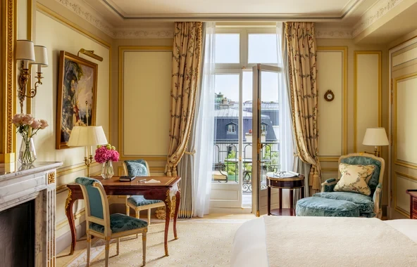 Aydınlık ve zarif bir şekilde dekore edilmiş lüks bir otel odasında, dışarıya açılan büyük Fransız balkon kapısından gün ışığı içeri süzülürken, odanın sol tarafında pembe çiçeklerle süslenmiş ahşap bir çalışma masası, karşısında teal renkli bir sandalye ve sağ tarafta uyumlu bir dinlenme koltuğu ile gold tonlarında şık perdeler görülüyor.