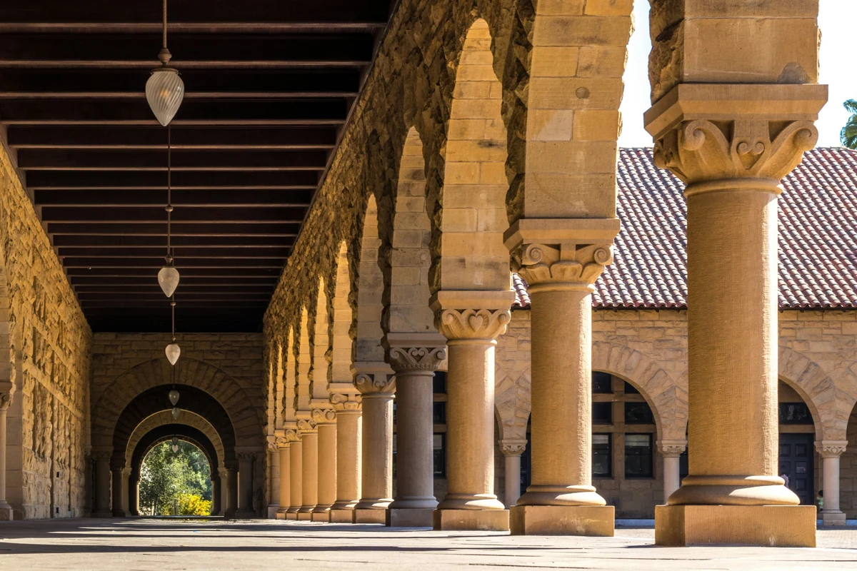Stanford Üniversitesi'nin karakteristik kahverengi taş binaları, kemerli geçitleri, yemyeşil çimlerle kaplı geniş avlusu ve sıralı palmiye ağaçlarıyla güneşli bir günde çekilmiş, huzurlu ve ilham verici akademik ortamını sergileyen panoramik bir görünüm.