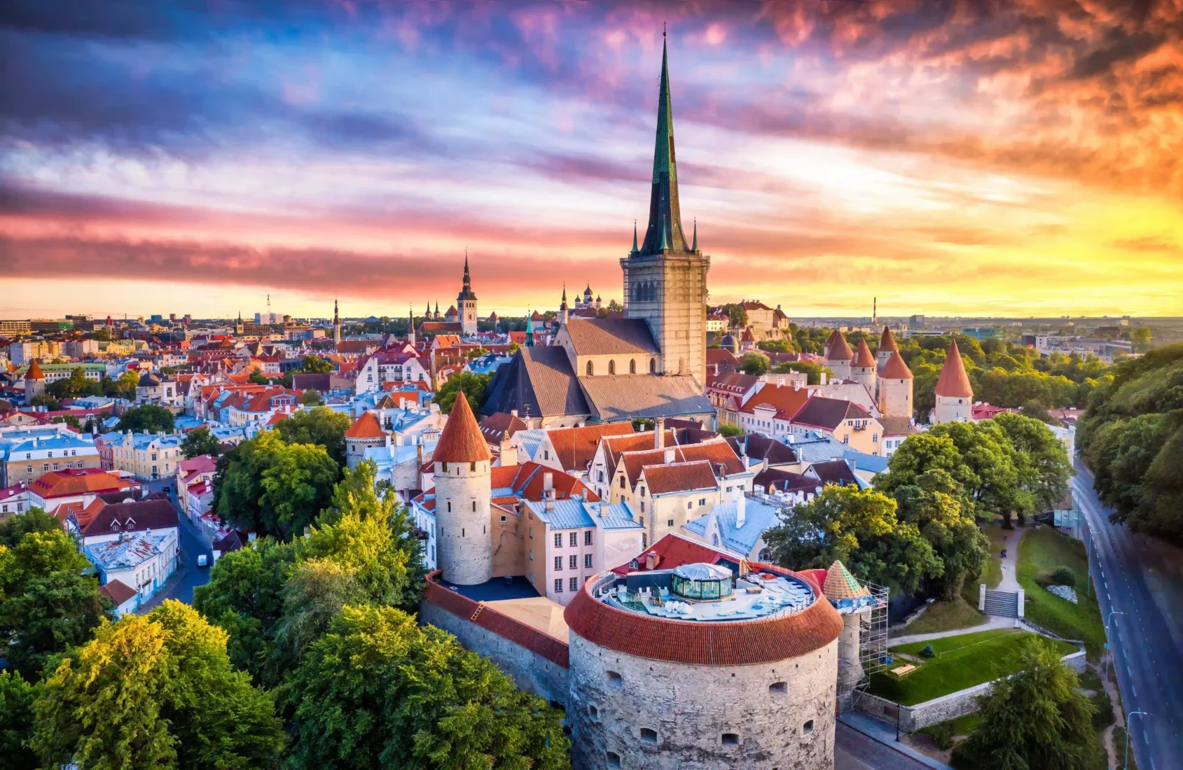 Tallinn'in Tarihi Eski Şehir bölgesindeki Arnavut kaldırımlı bir sokakta, kırmızı ve sarı cepheli, geleneksel mimariye sahip binaların sıralandığı, ileride sivri çatılı bir kulenin de seçilebildiği kemerli bir geçitten geçen ve şehrin canlı atmosferini yansıtan insanlar.