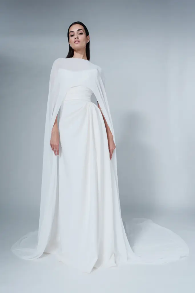 Minimal cape detaylı, drapeli beyaz uzun gelinlik. Modern ve heykelsi çizgileriyle öne çıkan zarif bridal siluet, 2026 gelinlik trendlerinin sofistike yorumunu yansıtıyor.