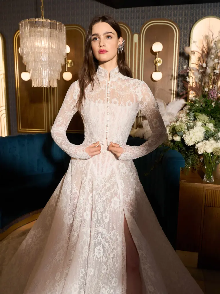 Yüksek yakalı, tamamen dantel işlemeli uzun kollu gelinlik giyen model; romantik A-line silüet ve yırtmaç detayıyla zarif bir bridal görünüm.
