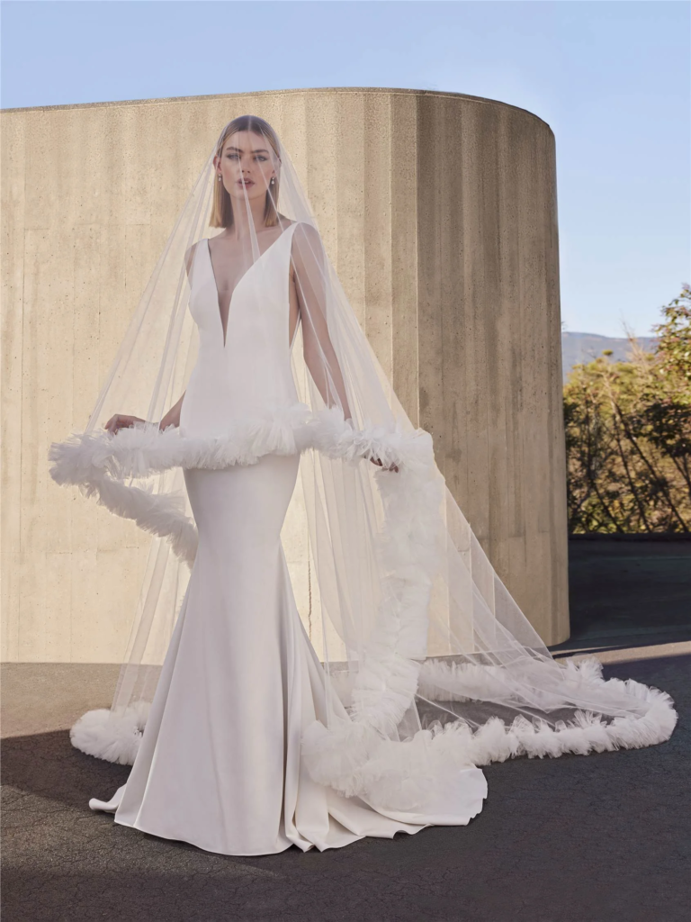 Derin V yakalı, sade ve heykelsi bir gelinlik giyen model; dramatik tül ve volan detaylı uzun duvakla modern bir bridal silüet sergiliyor.