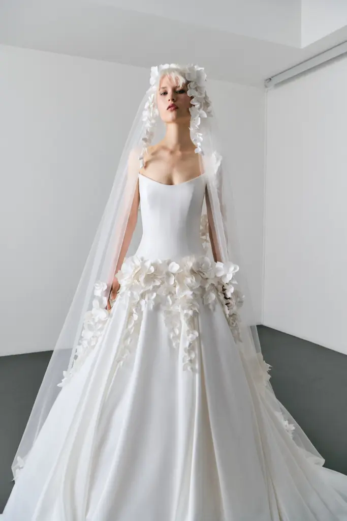 Üç boyutlu çiçek aplikeleriyle süslenmiş modern prenses kesim gelinlik ve floral detaylı uzun tül duvak. Minimal korse üstü ve heykelsi etek formuyla 2026 gelinlik trendlerini yansıtan romantik ve sofistike bridal görünüm.