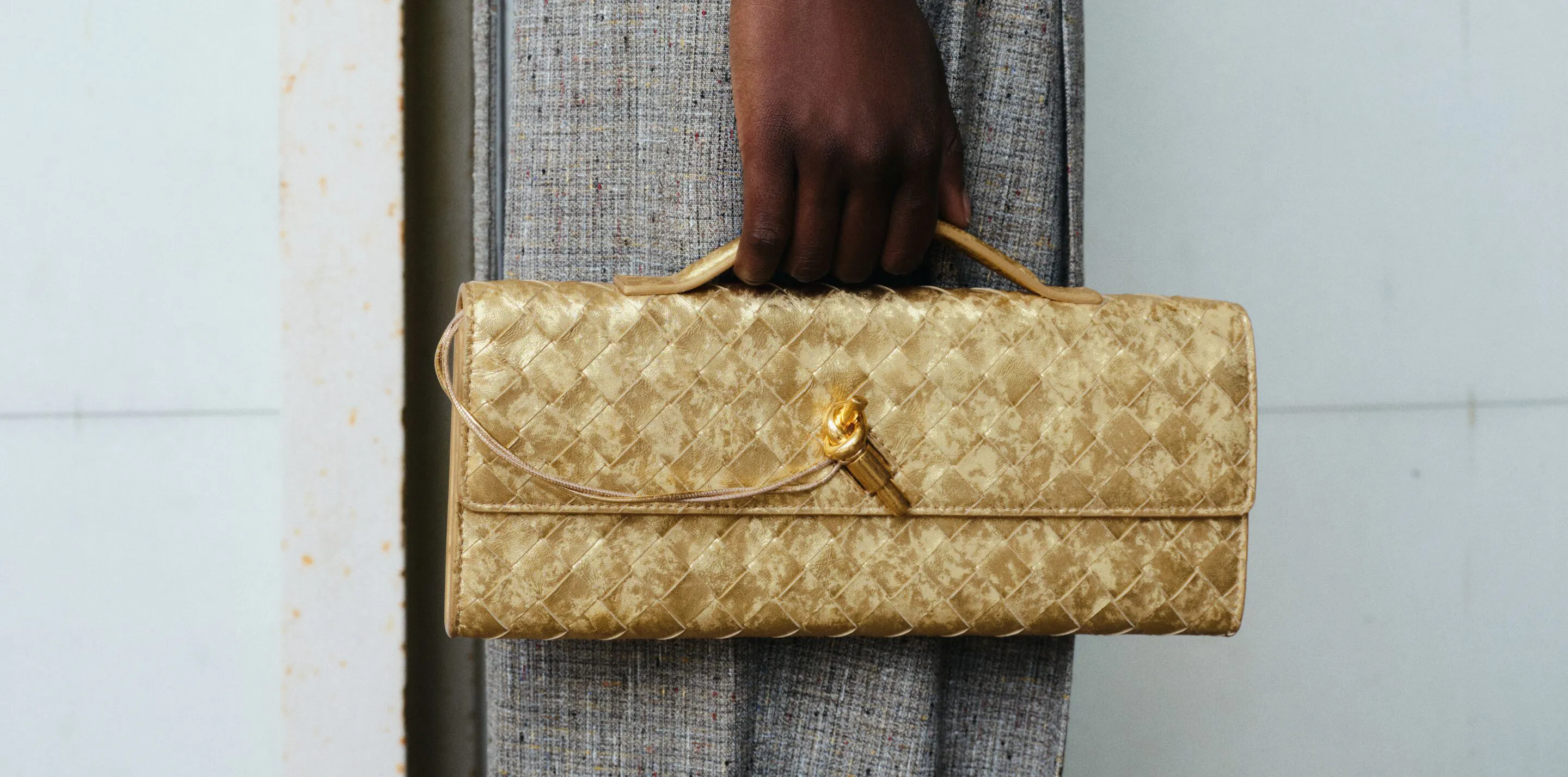 Altın tonlarında örgü dokulu lüks clutch çanta, bir modelin elinde minimalist arka plan önünde sergileniyor; 2026 moda trendlerinde öne çıkan dokulu çanta tasarımı.