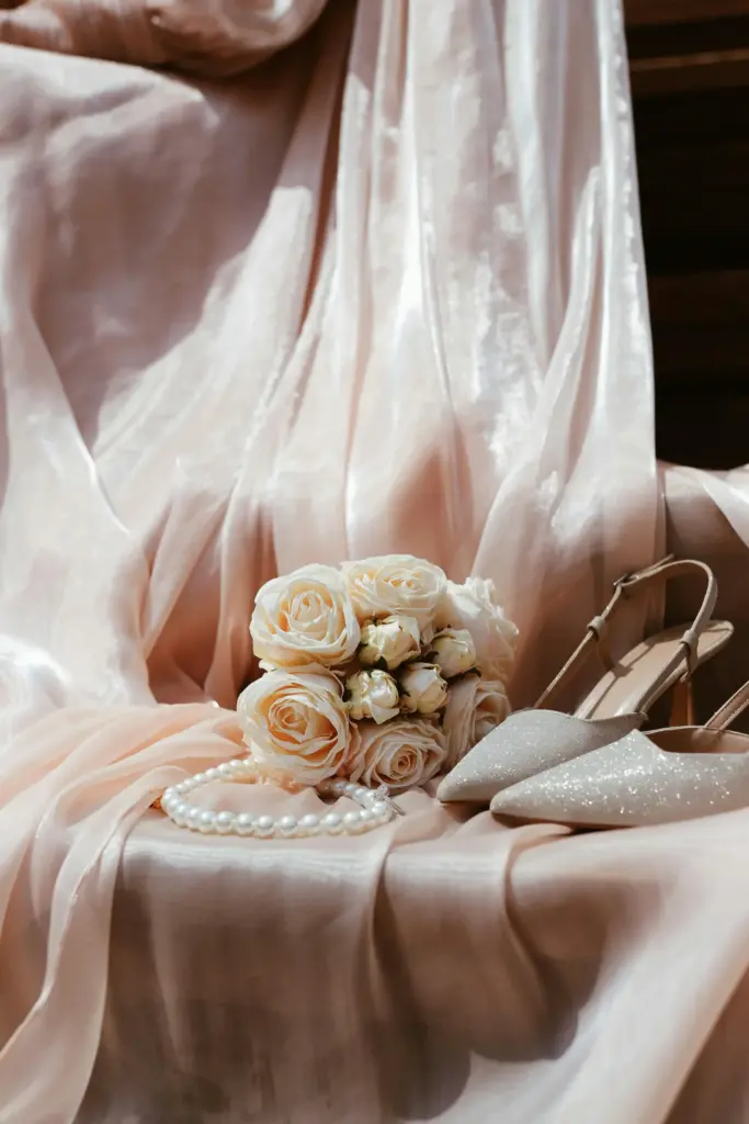 Şampanya tonlarında saten kumaş üzerinde beyaz gül buketi, ışıltılı gelin ayakkabıları ve zarif inci kolyesiyle romantik gelin aksesuarları yakın çekim still life fotoğrafı.