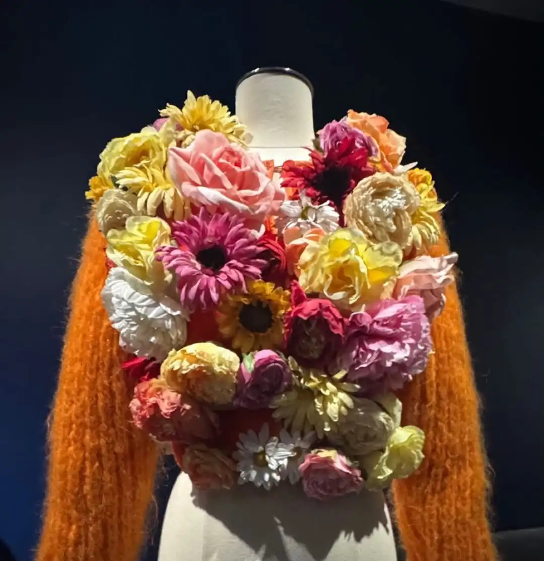 Canlı renklerde çiçek aplikeleriyle kaplanmış, heykelsi formda couture tasarım; doğa, el işçiliği ve yüksek modanın kesiştiği sanatsal sergi parçası.