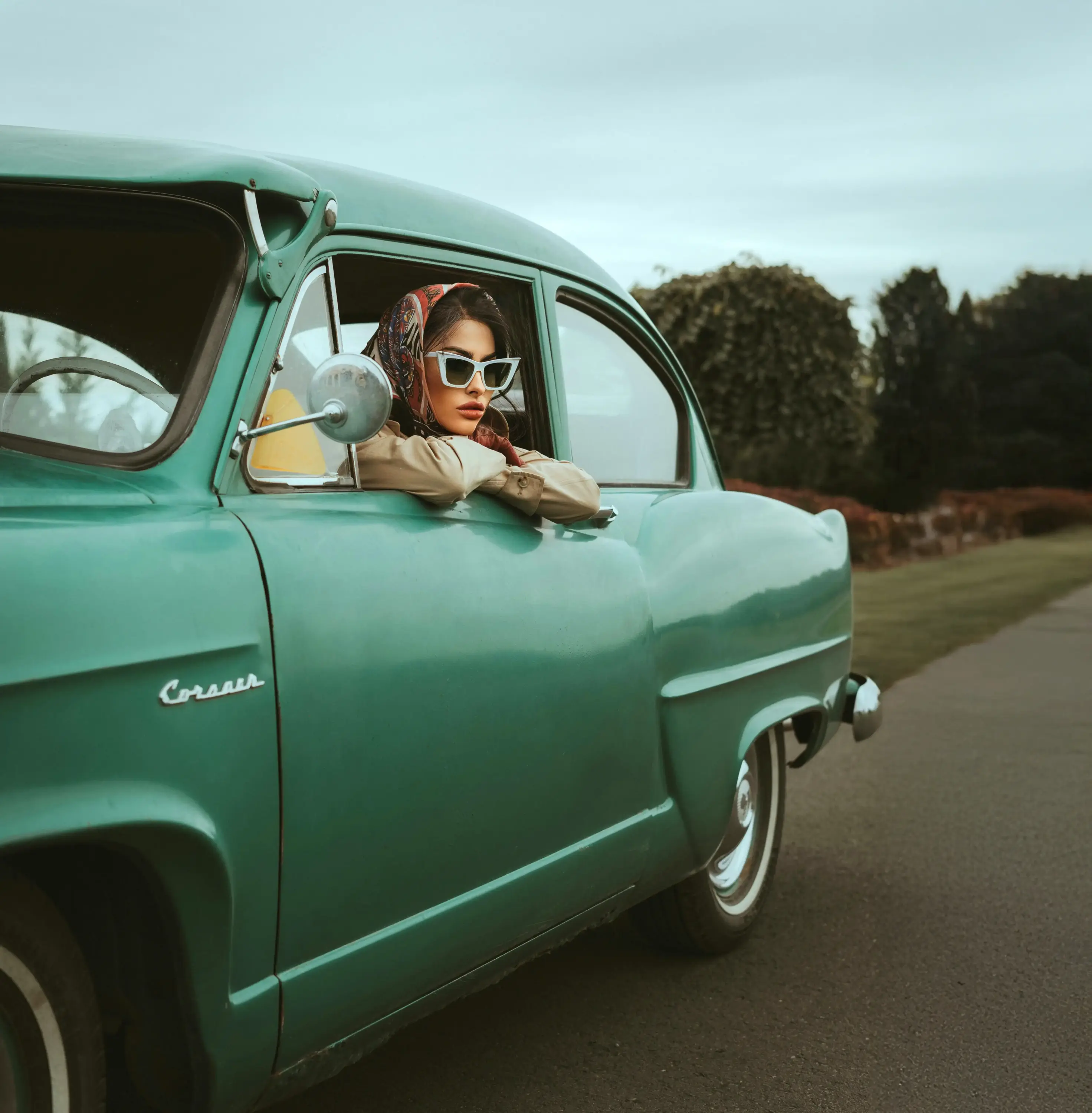 Vintage yeşil bir otomobilin penceresinden dışarı bakan şal ve güneş gözlüğü takmış modelin retro estetikte çekilmiş moda fotoğrafı.