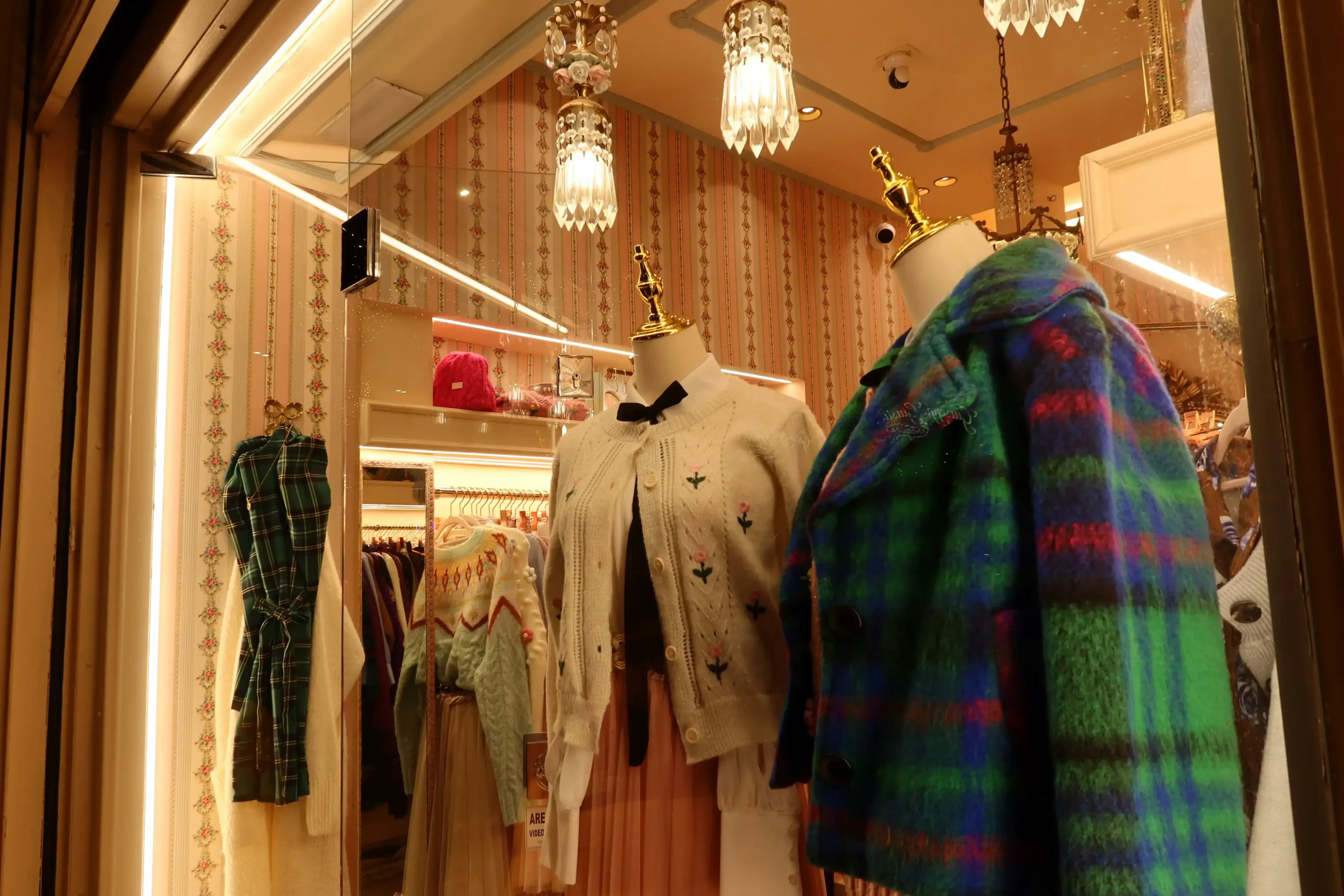 Vintage esintili butik vitrininde pastel örgü hırkalar ve ekose desenli renkli kabanların sergilendiği sıcak atmosferli moda vitrini.