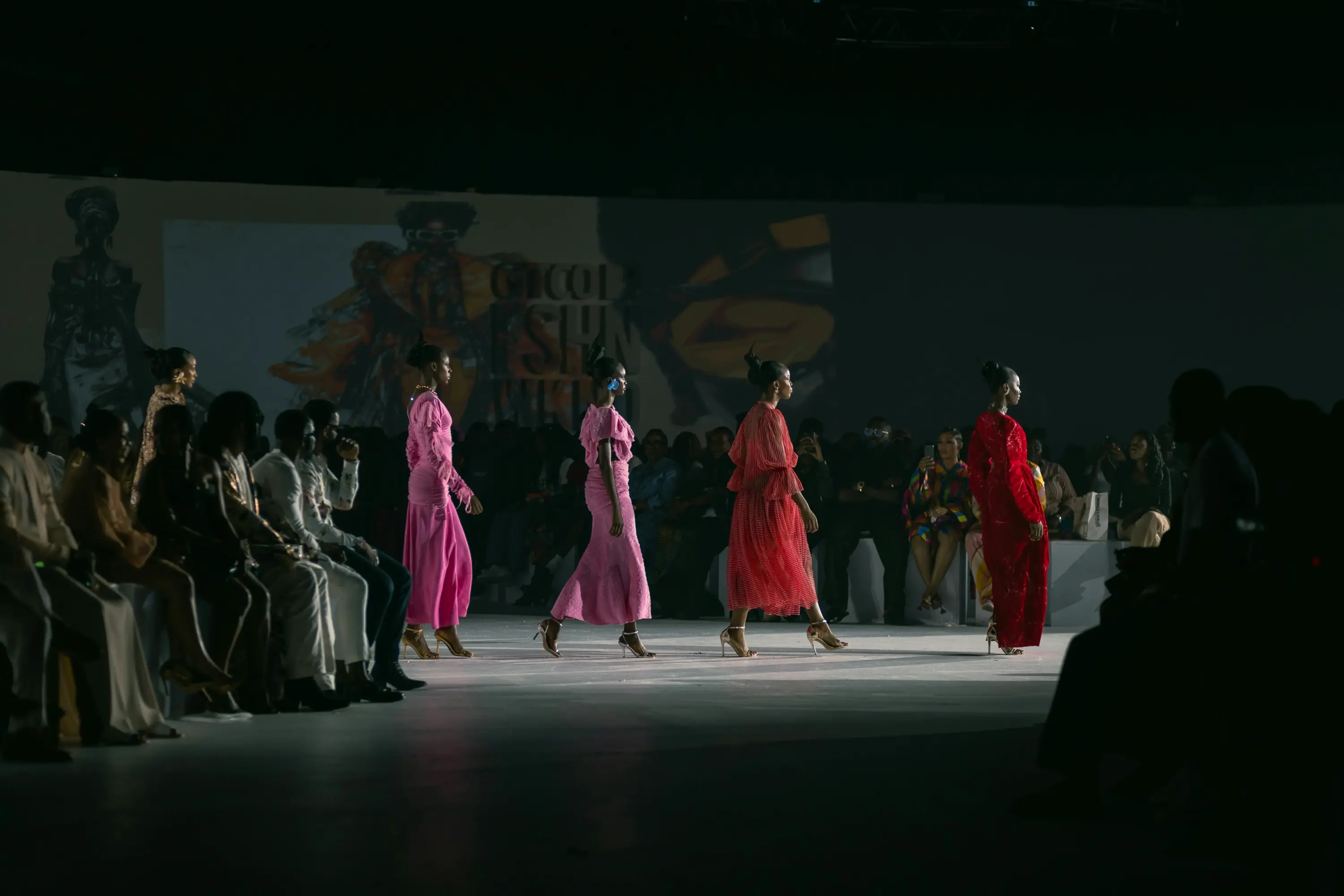 Podyumda pembe ve kırmızı tonlarda tasarımlar sergileyen modellerin yürüdüğü, kalabalık izleyici kitlesiyle gerçekleşen bir moda defilesi sahnesi.