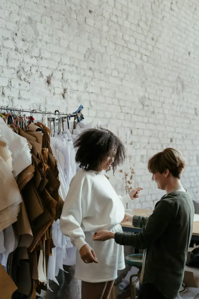 Bir tasarımcının atölyede model üzerinde prova yaptığı, kumaş numuneleriyle çevrili yaratıcı moda üretim sürecini gösteren çalışma sahnesi.