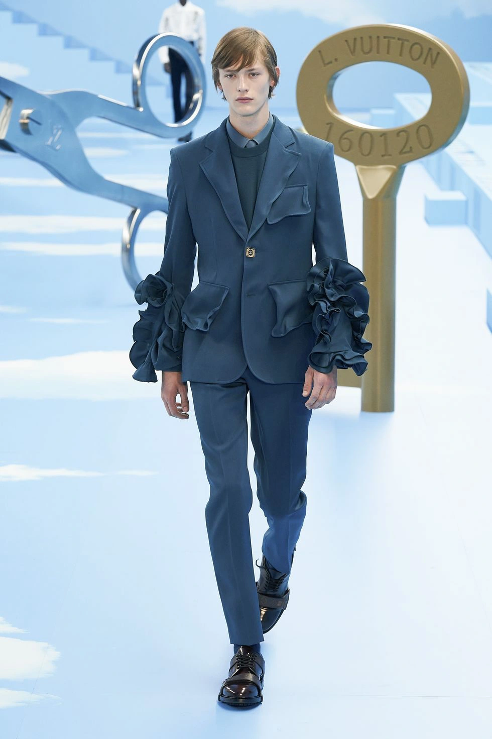 Louis Vuitton erkek podyumunda volanlı kol detaylarına sahip mavi takım elbise giyen model; yaratıcı siluetlerle öne çıkan modern erkek moda tasarımı.